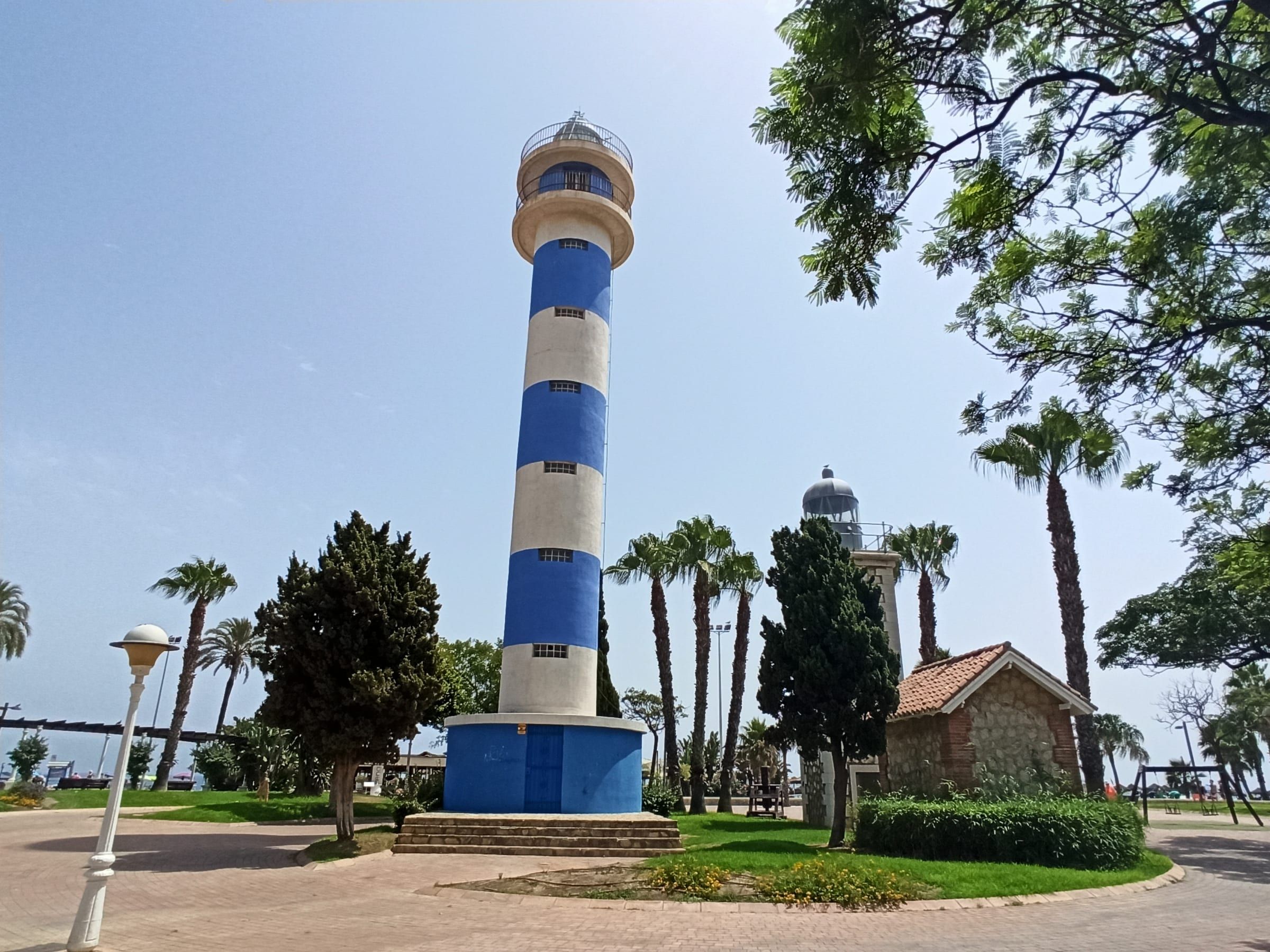 Faro de Torre del Mar, torre cilíndrica de franjas azules y blancas, con el antiguo faro de piedra detrás entre palmeras