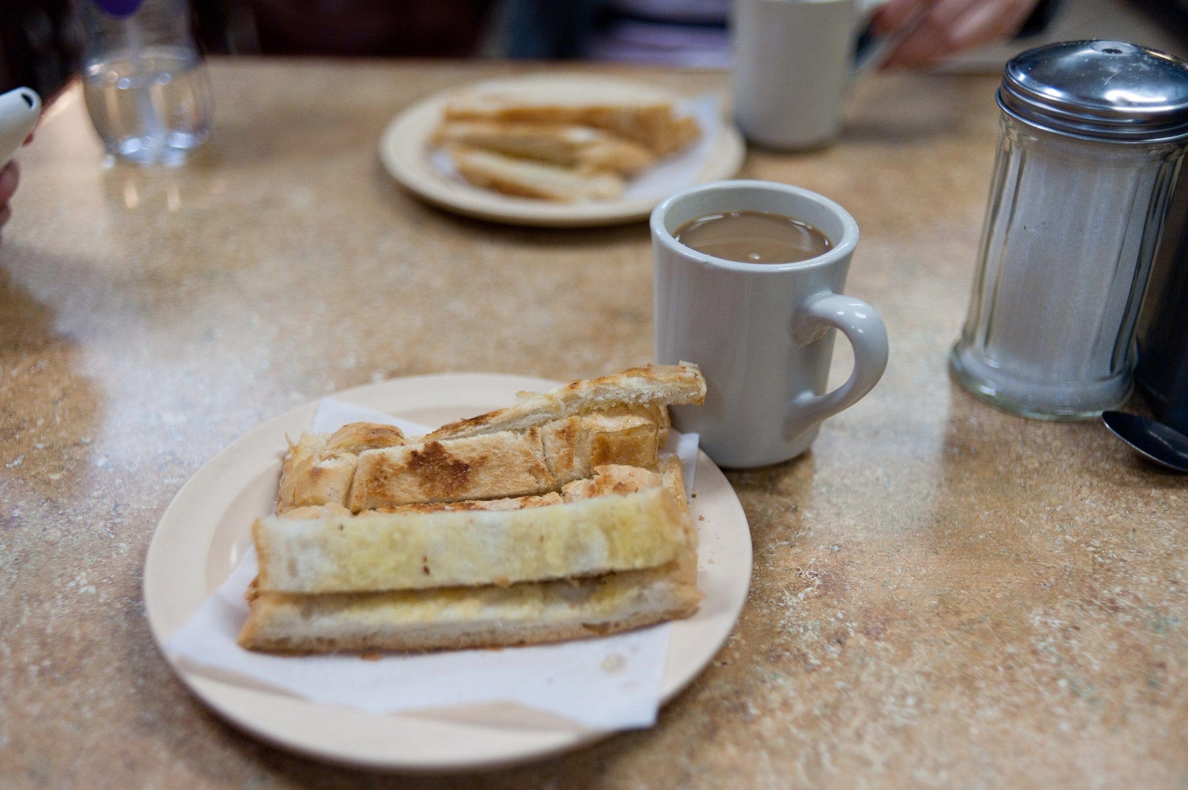 Desayuno en la barra de una cafetería, con una taza de café con leche, tostadas en el plato y el azucarero de cristal al lado