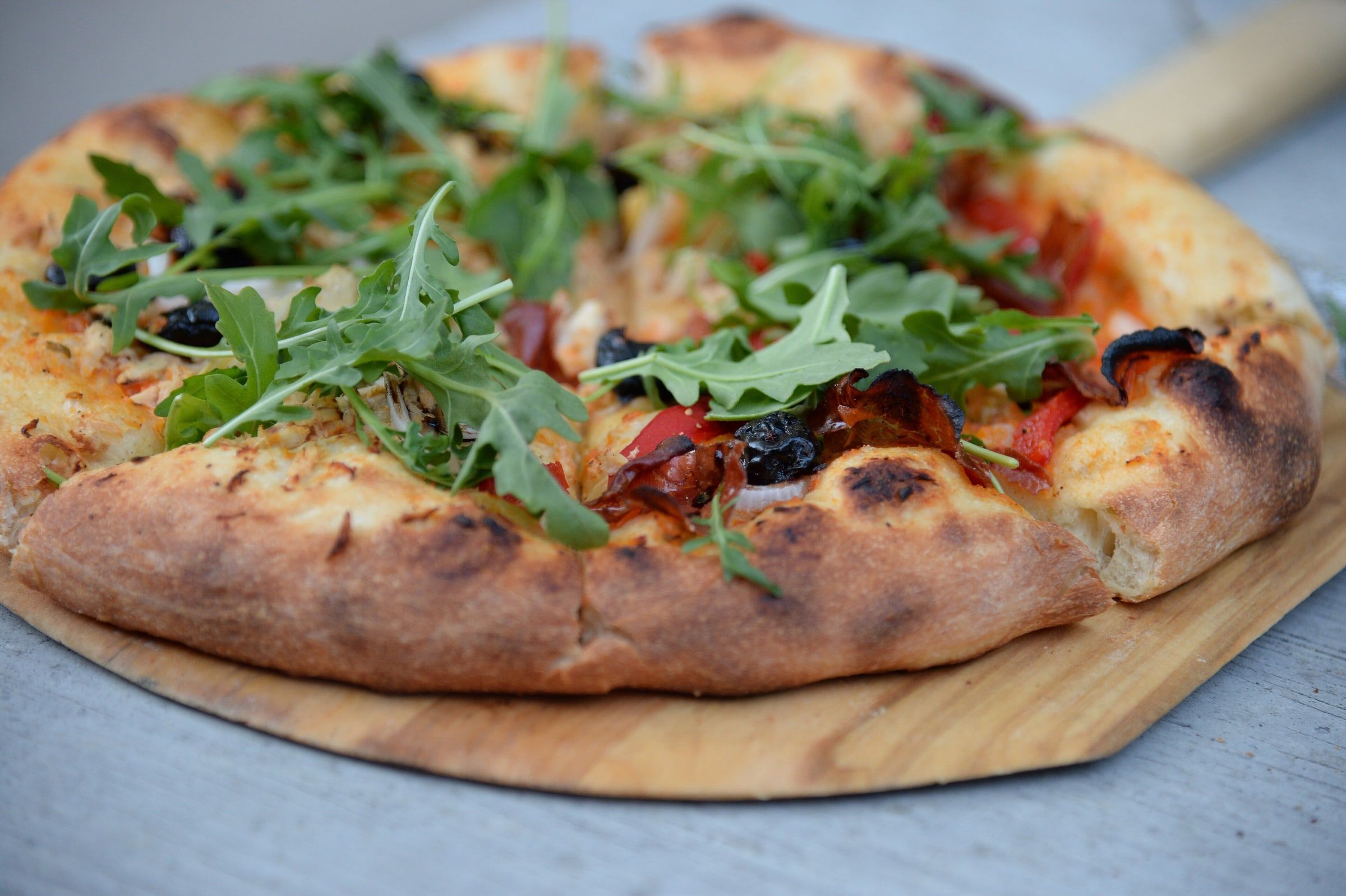 Pizza de masa fina con bordes tostados al horno de leña, cubierta de rúcula, aceitunas negras y tomate, sobre una pala de madera