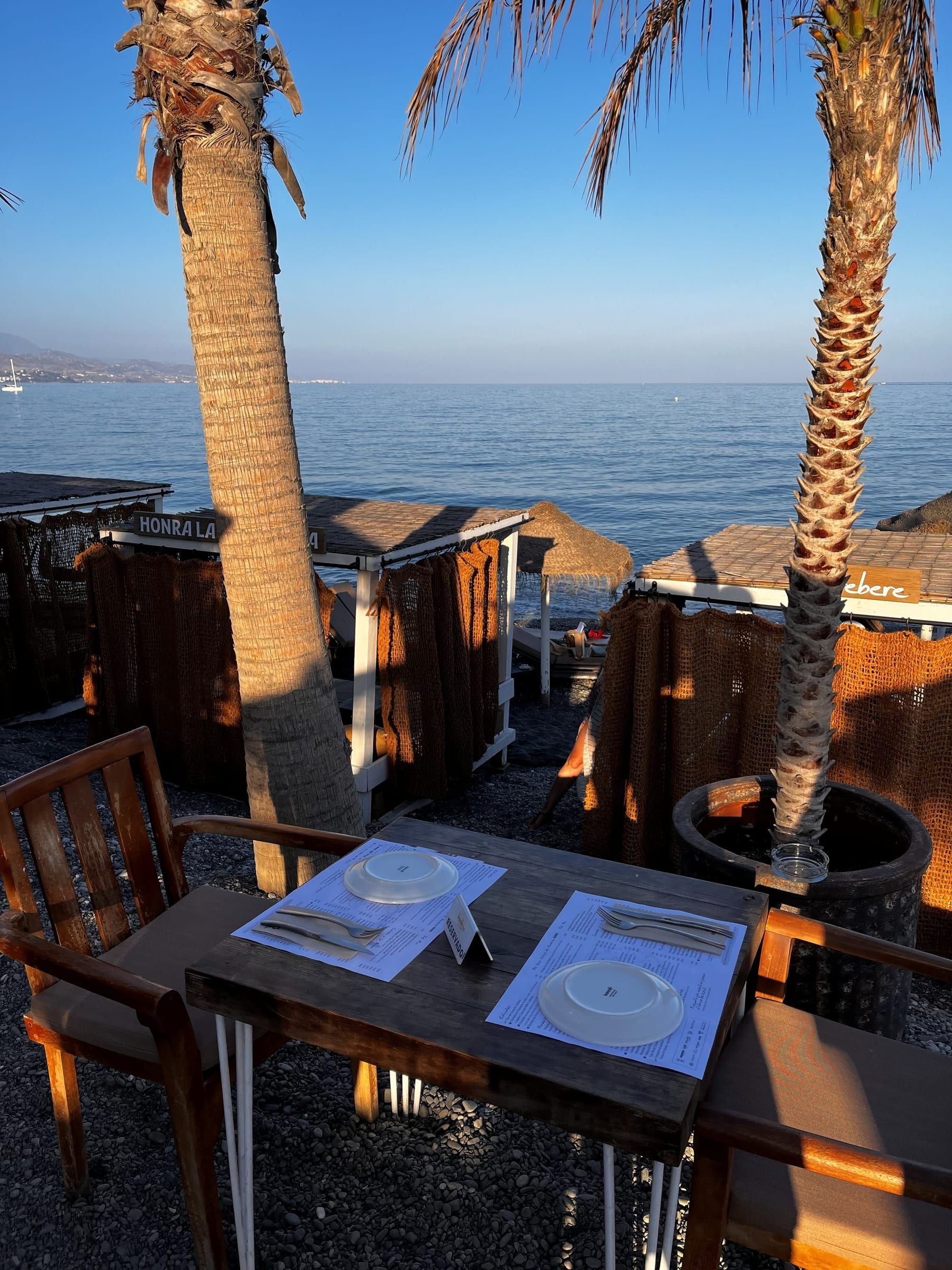 Mesa puesta junto al mar en un chiringuito del paseo marítimo de Torre del Mar al atardecer, con palmeras y las montañas de la Axarquía al fondo