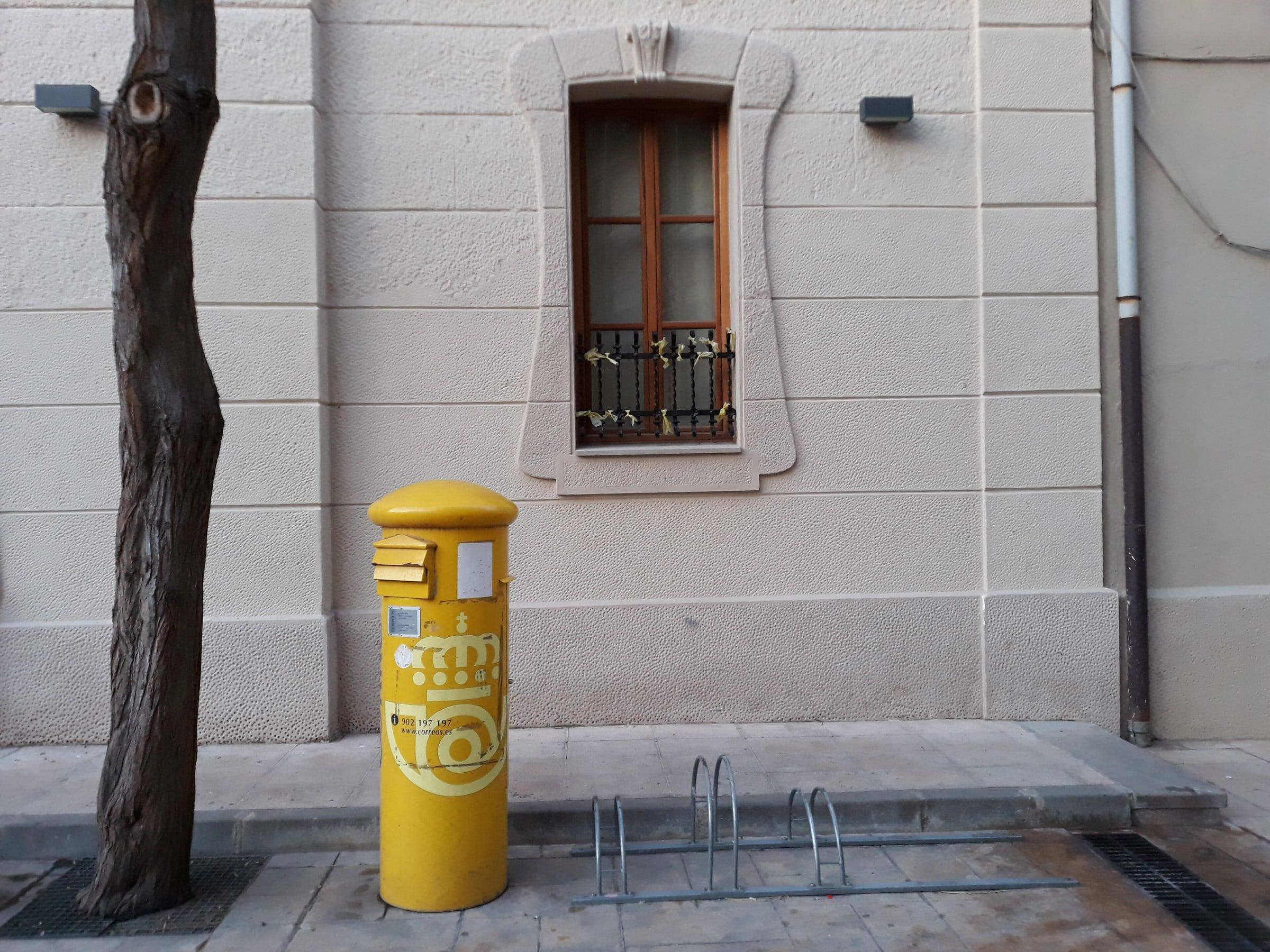 Buzón amarillo de Correos en una plaza, junto a la fachada blanca de un edificio con ventana y balcón