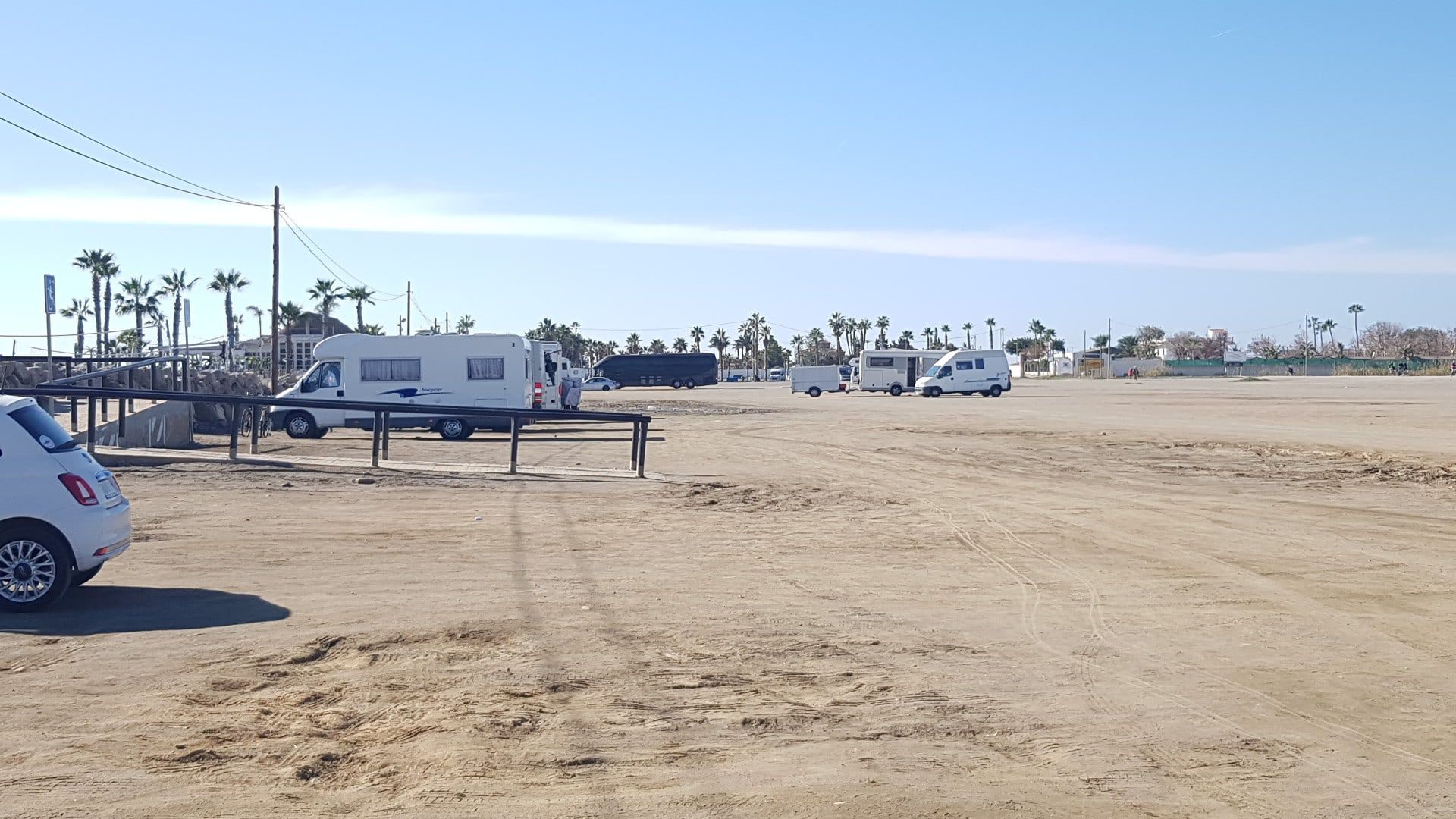 Explanada de tierra junto a la playa de Torre del Mar usada como aparcamiento, con coches y autocaravanas y palmeras al fondo